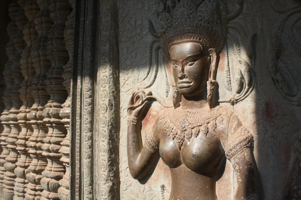 An apsara carved in stone relief at Angkor Wat, in a tall headdress with one hand raised, beside a pillar of turned stone.