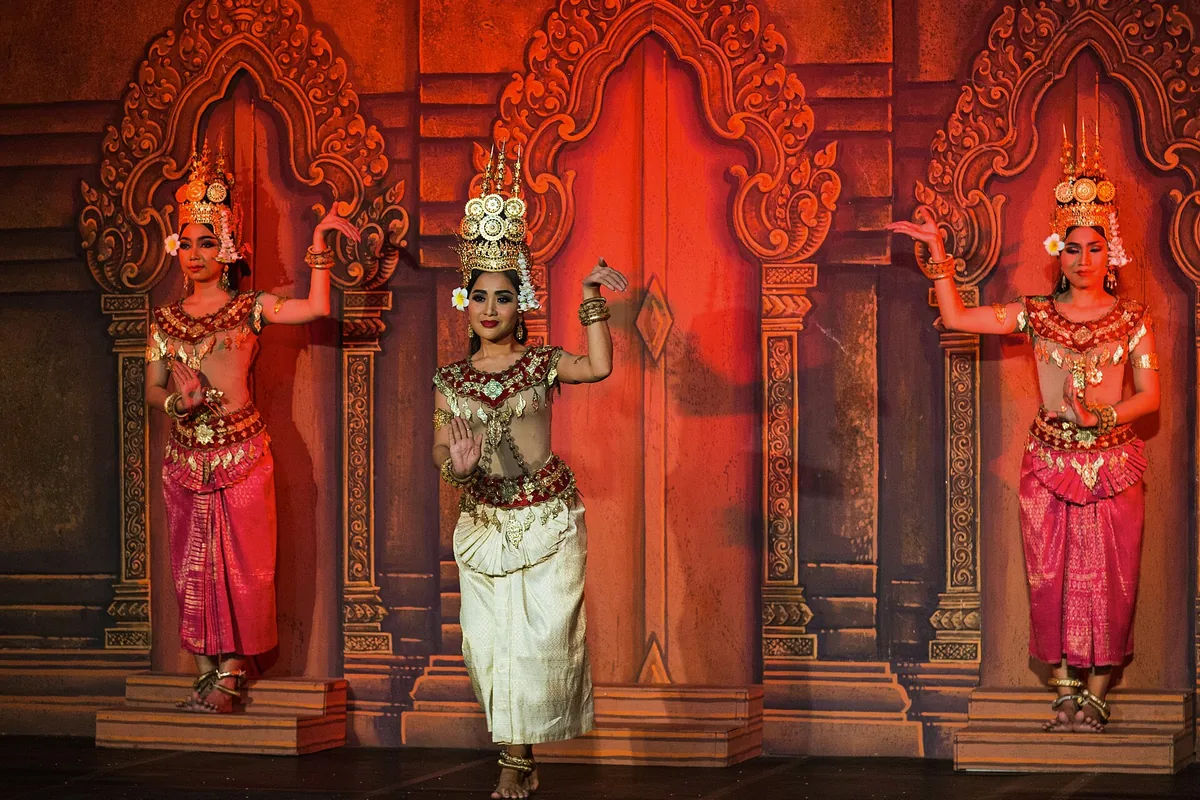 Three apsara dancers in front of a carved temple-style backdrop, the centre dancer in a white skirt-cloth and a tall gold crown, her flanking dancers in magenta.