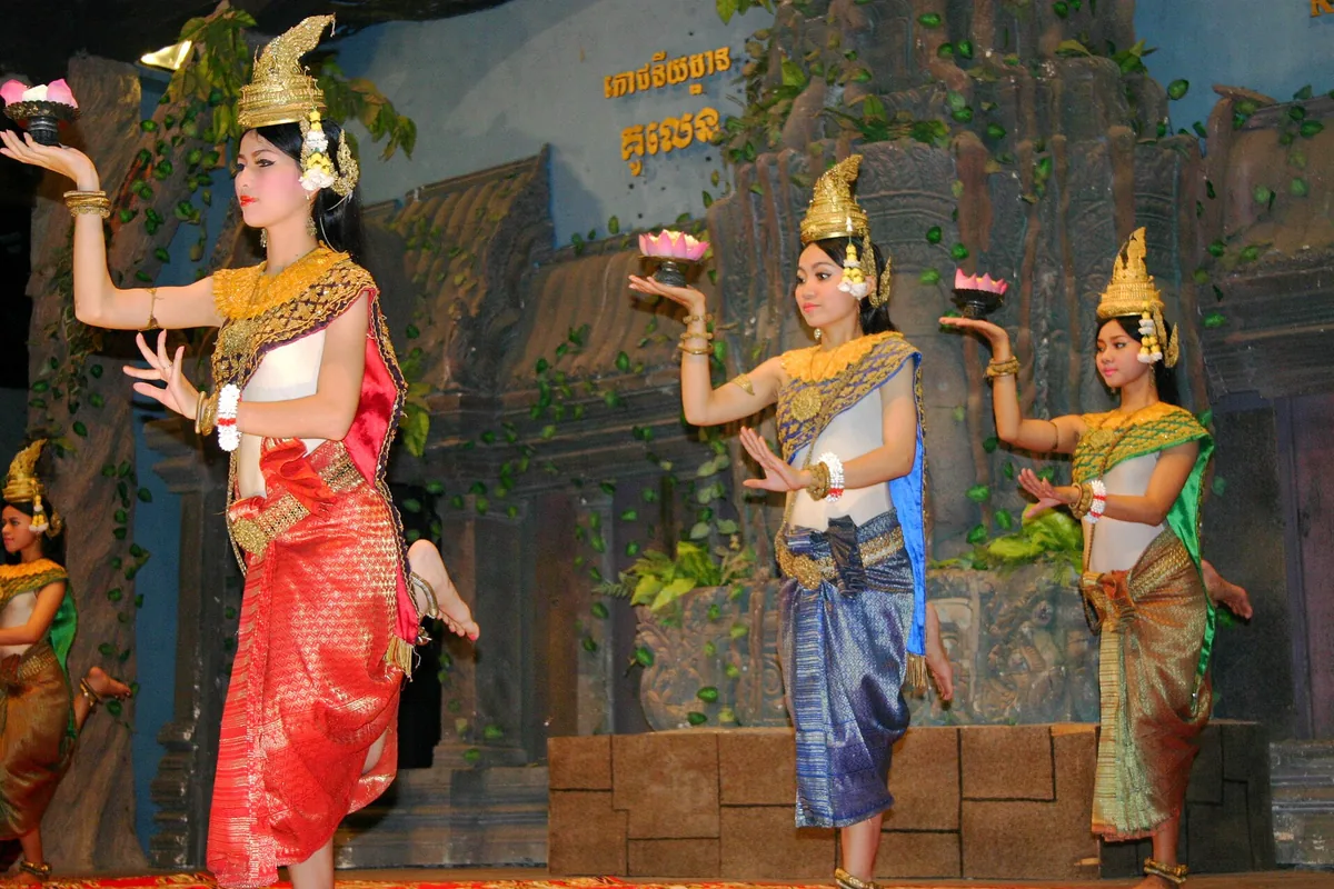 Three apsara dancers holding pink lotus flowers, in gold crowns and red, blue and green silk, on a stage set with stone blocks and vines.