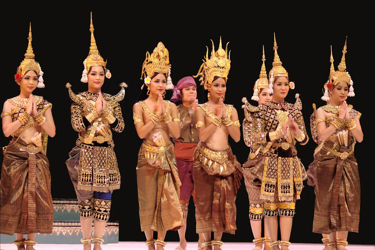 Dancers of the Royal Ballet of Cambodia at a curtain call in Paris, hands together, in gold spired crowns and brocade costumes on a dark stage.