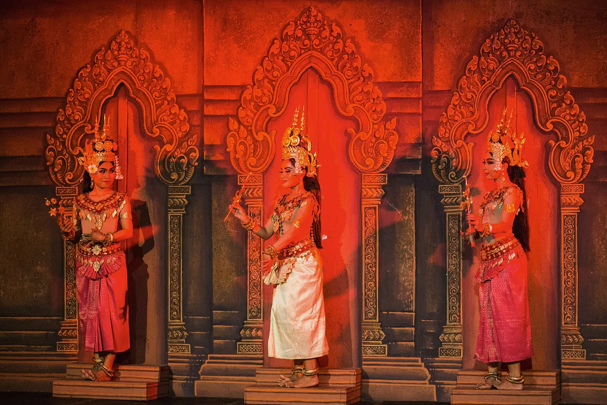 Three apsara dancers in gold crowns, each standing in front of a carved arch in a red-lit temple-style backdrop, the centre dancer in white.