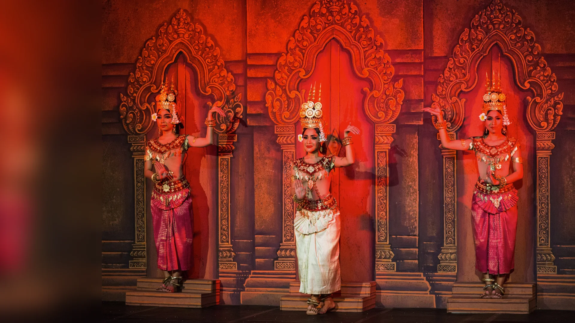 Three apsara dancers in gold crowns and silk before a carved temple-style backdrop lit red, the dancer in the centre dressed in white.